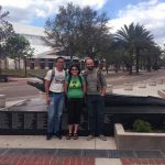 Three people standing in front of a bronze alligator statue on a university campus, with trees and a walkway in the background.