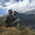 Two men sit on a grassy mountainside with snow-capped peaks in the background, smiling and posing for the camera under a partly cloudy sky.