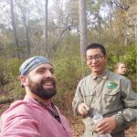 Two men are outdoors; one in a pink shirt takes a selfie while the other in a green uniform holds a water bottle. A woman is in the background surrounded by trees and foliage.