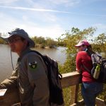 Two people with backpacks stand on a wooden platform overlooking a body of water and vegetation. One person, wearing a wide-brimmed hat, holds a paper while the other wears a pink cap.