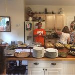 A group of people gathered near a kitchen island containing various dishes and plates. One person at the center is speaking, while others are looking on and engaging in conversation.