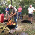 A man grills food over a flaming barbecue while three men and a dog are nearby. The scene is set in a grassy, wooded yard during the daytime.