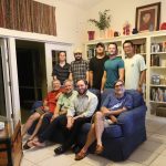 A group of nine men of varying ages pose together in a living room. Some are seated on couches while others stand behind. The room has bookshelves, decorative items, and a glass door.