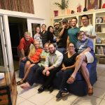 A group of thirteen people smile for a photo in a living room. Most are seated on couches, while others stand behind them. Bookshelves and home decor are visible in the background.