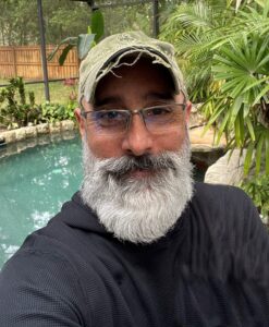 A man with a gray beard, glasses, and a green cap takes a selfie outdoors in front of a pool and tropical plants.