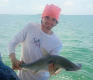 A man wearing a pink headscarf and white long-sleeved shirt holds a hammerhead shark over the water.