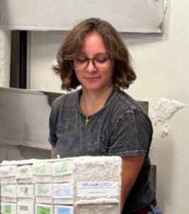 A person wearing glasses and a gray t-shirt stands behind a white, textured rectangular object.