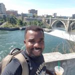 A person holds an iced coffee while standing in front of a river and a bridge, with buildings in the background, on a sunny day.