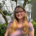 A person with long hair and glasses wearing a purple shirt stands outside in front of a window, smiling at the camera. There are green shrubs and a small tree in the background.