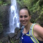 A woman in a blue tank top, with a hydration pack, smiles while standing in front of a waterfall surrounded by lush greenery.