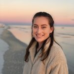 A woman with long brown hair, wearing a beige fleece jacket, stands on a beach at sunset, smiling at the camera.