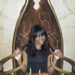 A woman with long dark hair sits and smiles on a large, elaborate wooden chair with curved armrests.