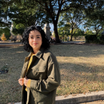 A person with curly hair and an olive green jacket stands outside on a sunny day, with trees and a park in the background.