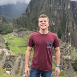 A man wearing a maroon t-shirt and jeans stands on a grassy area with Machu Picchu ruins and mountains in the background.