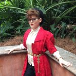Person with short curly hair and glasses wears a red coat with a gold belt, leaning against a stone ledge with a background of lush green plants.