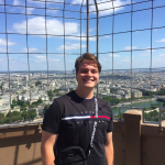 A person smiles while standing on an observation deck with a cityscape and blue sky in the background.