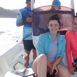 A woman in a light blue shirt and shorts smiles while sitting on a boat. She is holding a black object, and a person in a red shirt is partially visible next to her. Another person stands in the background.