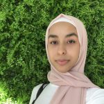 A person wearing a light pink hijab and a white top stands in front of a lush green leafy backdrop.