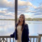 A woman in a white outfit and dark jacket stands smiling on a wooden deck overlooking a calm lake under a partly cloudy sky.