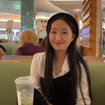 A person with long black hair, wearing a black beret and white shirt, sits at a table in a dining area with a plastic cup in front of them.