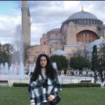 A person stands in front of the Hagia Sophia with a fountain in the background, wearing a plaid shirt and carrying a bag. The sky is partly cloudy.