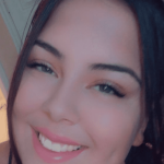 A woman with long dark hair smiles at the camera, showing her teeth. She is indoors and wearing a sleeveless top.