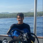A person in a wetsuit smiles while standing on a boat with scuba diving equipment in front. The backdrop includes a body of water and a distant shoreline.