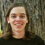 A young person with long brown hair and a black shirt smiles in front of a tree trunk.
