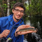 Person in blue jacket and glasses holding a small fish near a wooded water area, smiling at the camera. A fishing rod is also visible.
