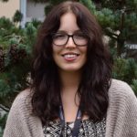 A woman with long curly hair, wearing glasses, a patterned top, and a beige cardigan, stands smiling outdoors in front of greenery.