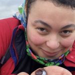 A person wearing a colorful headscarf and red jacket holds a small seashell and some sand in their hand, smiling at the camera.