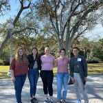 Five people stand outdoors on a sunlit day with trees and a light post in the background, posing for the photo.