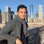 A man wearing a gray scarf and coat stands on a bridge with a city skyline in the background.