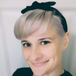 Person with short, light-colored hair and a black headband smiling at the camera. Wearing a black top and a necklace. Soft-focus background.