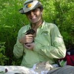 Person sitting in greenery, smiling, and holding a small animal. They wear a khaki hat and light green shirt.