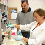 Two people working in a laboratory, one wearing a lab coat and gloves while operating equipment, the other observing.