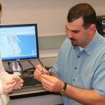 Two people are examining small objects in front of a computer displaying a map. They are in an office setting.