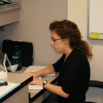 A person with curly hair sits at a desk, writing on paper with a laptop open, in front of a whiteboard.
