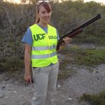 A person in a UCF Biology vest holds a shotgun outdoors in a grassy area.