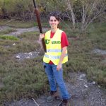 Person in a yellow safety vest and jeans holding a shotgun, standing outdoors on grassy terrain with trees in the background.
