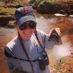 Person wearing a cap and sunglasses, holding a turtle by a pond. A pair of binoculars hangs around their neck.