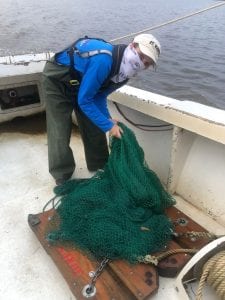 Person in outdoor gear adjusts a green net on a boat deck by the water.