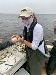 Person in fishing gear holds a small fish on a boat, with a tray of fish visible. Overcast sky and water in the background.