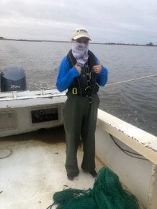 Person in a blue shirt, green waders, and face covering gives two thumbs up on a boat by the water.