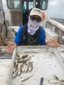 Person on a boat wearing a hat and bandana, shrugging, with a display of small fish on a tray in front of them.