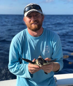 A person in a blue shirt stands on a boat holding a small sea turtle, with the ocean and blue sky visible in the background.