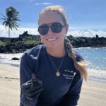 Person kneeling on a beach wearing sunglasses and holding a small turtle. Ocean and palm tree in the background.