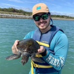 Person in a blue shirt and life vest holds a sea turtle on a boat, with a body of water and a rocky shore in the background.