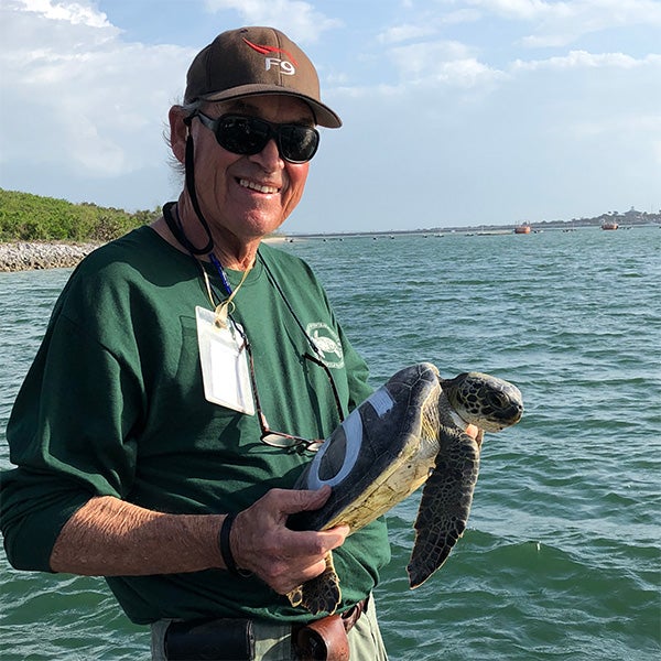 Man wearing a hat and sunglasses holds a sea turtle by the water, with a shoreline and cloudy sky in the background.