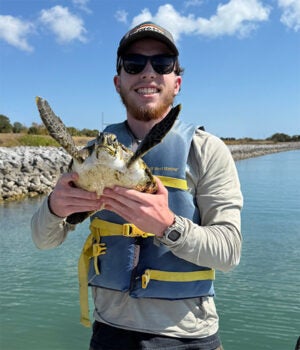 A person wearing sunglasses and a life jacket holds a small sea turtle while standing near water with rocks and trees in the background.